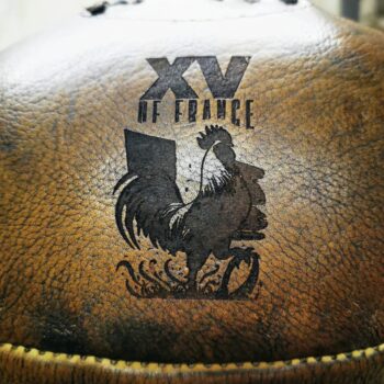 Ballon de rugby vintage en cuir havane, effet bois, avec gravure artisanale du coq et du logo XV de France. Photo de détail montrant la texture patinée, la couture traditionnelle et la personnalisation haut de gamme réalisée dans notre atelier. Idée cadeau rugby, customisation sur cuir et gravure personnalisée pour amateurs du XV de France et décor sportif authentique.
