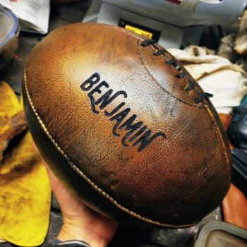 Ballon de rugby vintage en cuir havane, finition effet bois, personnalisé avec une gravure prénom “Benjamin”. Photo détaillée montrant la patine artisanale, les coutures traditionnelles et le rendu haut de gamme de la customisation. Idéal pour idée cadeau rugby, personnalisation sur cuir et créations inspirées des ballons en bois décoratifs.