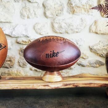 Ballon de football américain vintage Ben & Flo en cuir véritable avec logo Nike. Commande spéciale de la marque pour une édition limitée au style rétro. Patiné à la main dans notre atelier en France, ce ballon décoratif rend hommage à l’authenticité du sport américain et à l’élégance du cuir brun travaillé artisanalement.