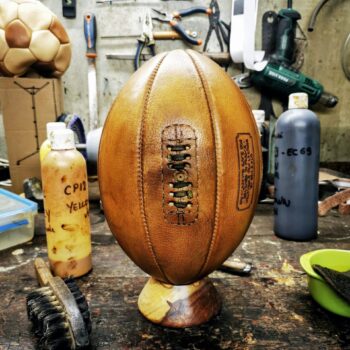 Ballon de rugby en cuir honey sur établi de l’atelier Ben & Flo, montrant le processus de patine artisanale à la cire et aux pigments naturels.
