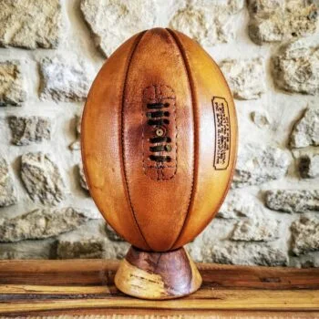Ballon de rugby vintage de collection à 8 panneaux en cuir véritable patiné à la main, présenté sur un socle en bois tourné. Réalisé artisanalement dans l’atelier Ben & Flo en France, ce ballon incarne le savoir-faire sportif traditionnel et l’élégance intemporelle du cuir naturel.