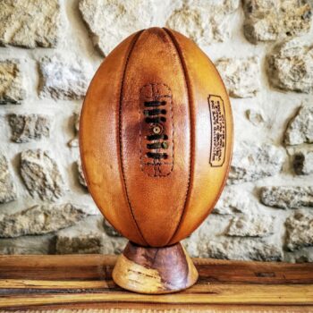 Ballon de rugby vintage de collection à 8 panneaux en cuir véritable patiné à la main, présenté sur un socle en bois tourné. Réalisé artisanalement dans l’atelier Ben & Flo en France, ce ballon incarne le savoir-faire sportif traditionnel et l’élégance intemporelle du cuir naturel.