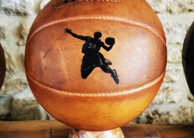 Ballon de basket vintage en cuir avec une gravure de Kobe Bryant en plein dunk, posé sur un socle en bois, contre un mur en pierre