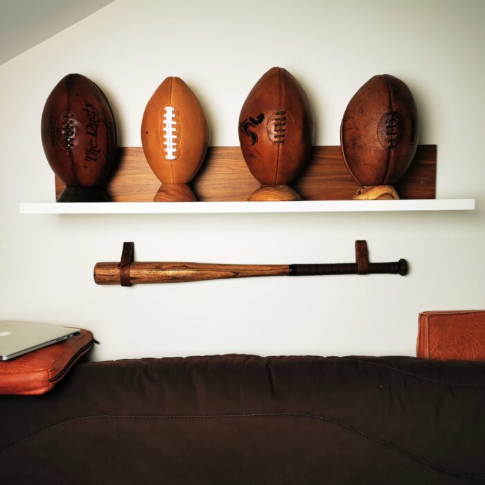 Décoration murale vintage avec une batte de baseball suspendue sur un support mural en cuir sous une étagère de ballons en cuir.