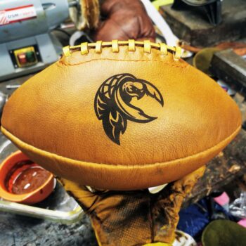 Ballon de foot américain en cuir patiné dans notre atelier, décoré d’un logo tortue tribal gravé au laser. Modèle vintage idéal pour la décoration football américain ou un cadeau original pour fan de football US. Vue de près sur la couture, la patine miel et l’ambiance artisanale de l’atelier, parfait pour les amateurs d’objets sportifs authentiques et faits main.